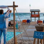 Mariage à la plage : couple ajuste des rubans sur arche en bois, plage coucher de soleil